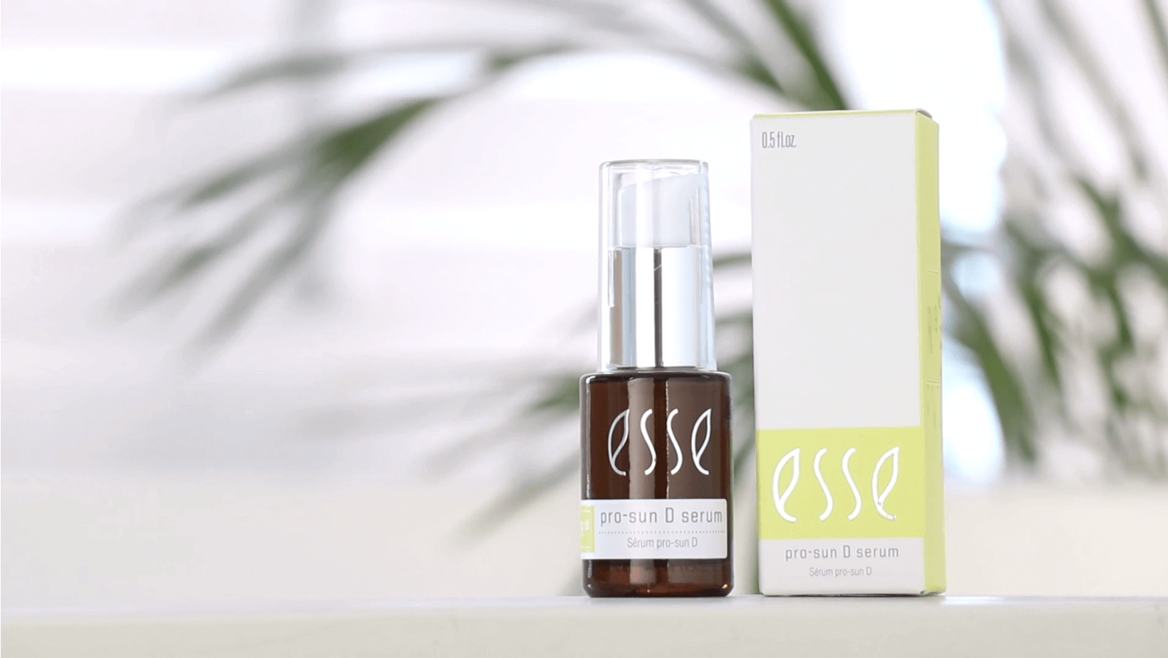 A bottle of Esse Pro-Sun D Serum is displayed next to its matching box. The bottle features a dark color with a white label, topped with a silver and clear cap. The box is light-colored and accented with green details. A blurred plant serves as the backdrop.