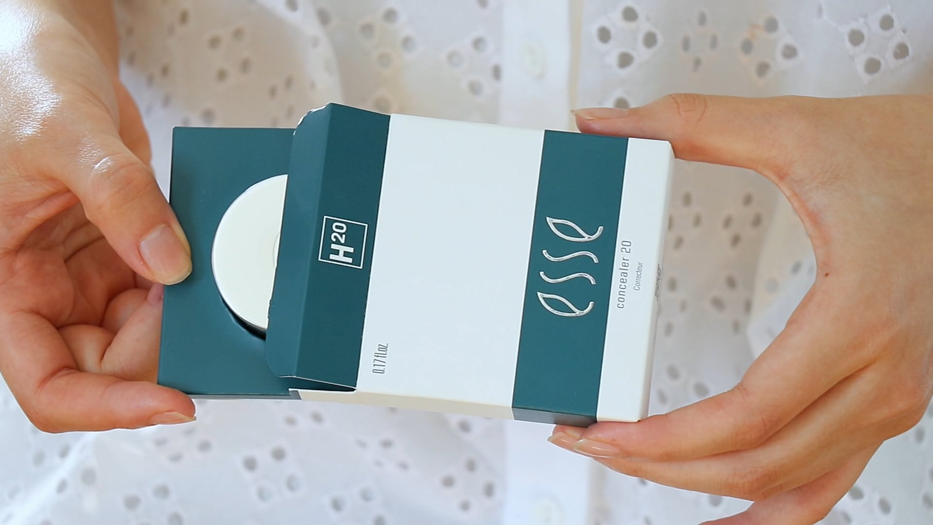 A person is holding a rectangular cosmetic box with a green and white color scheme. The box, labeled "H2O" and "Esse Concealer," is partially open, revealing a round white container inside. The background features a white, eyelet-patterned fabric.
